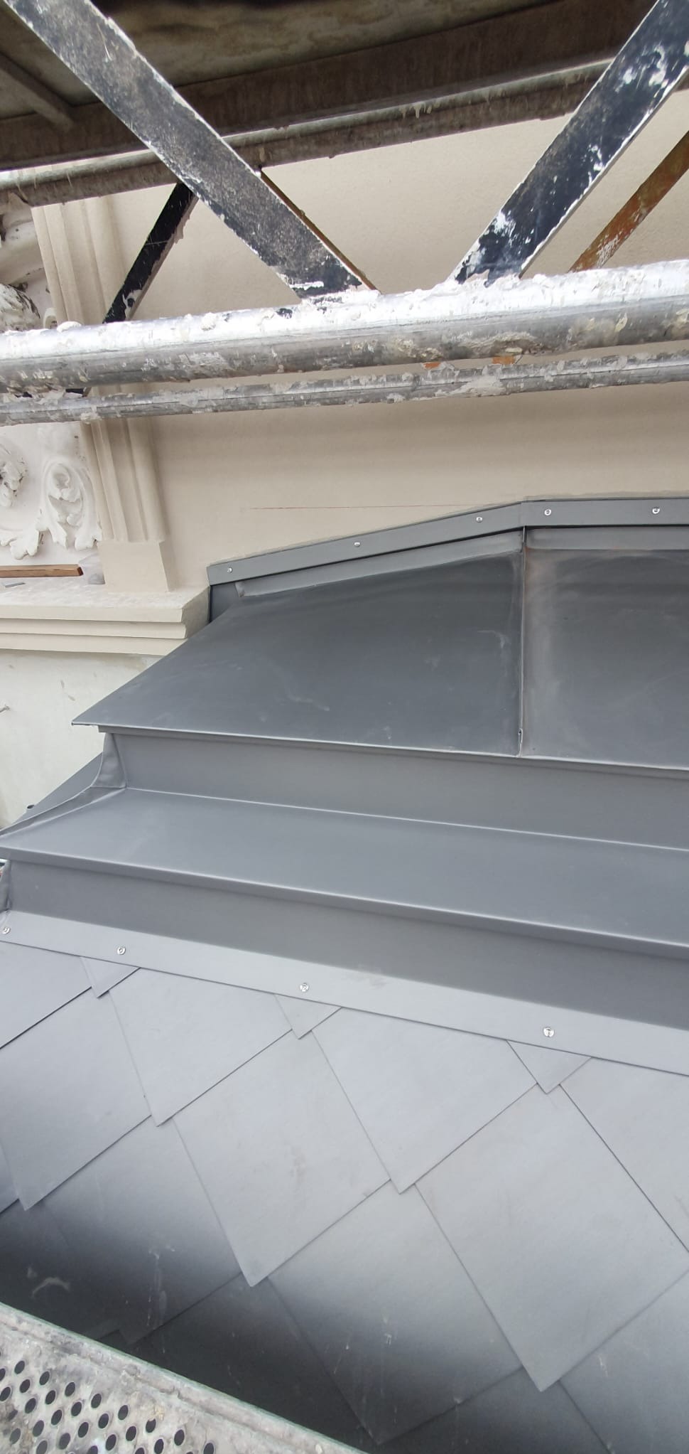 Szary dach z blachy ułożony w łuskę, widoczny z rusztowania. Detal obróbki blacharskiej przy ścianie budynku z elementami dekoracyjnymi.