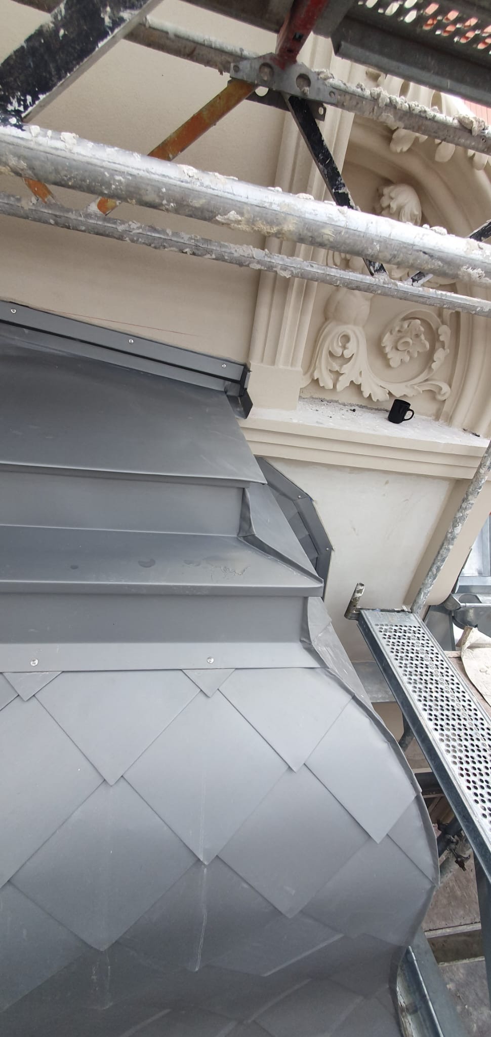 Szare, romboidalne płytki na dachu budynku z dekoracyjną fasadą, widoczne rusztowanie i czarny kubek na gzymsie, sugerujące przerwę w pracach.