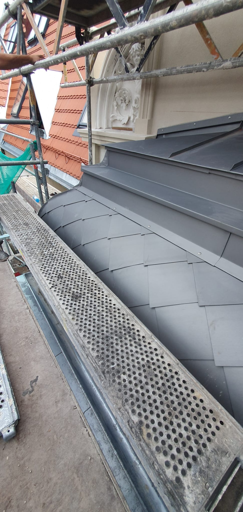 Szary dach z blachy ułożonej w romby, widoczny z rusztowania. W tle czerwona dachówka i ozdobny detal architektoniczny.