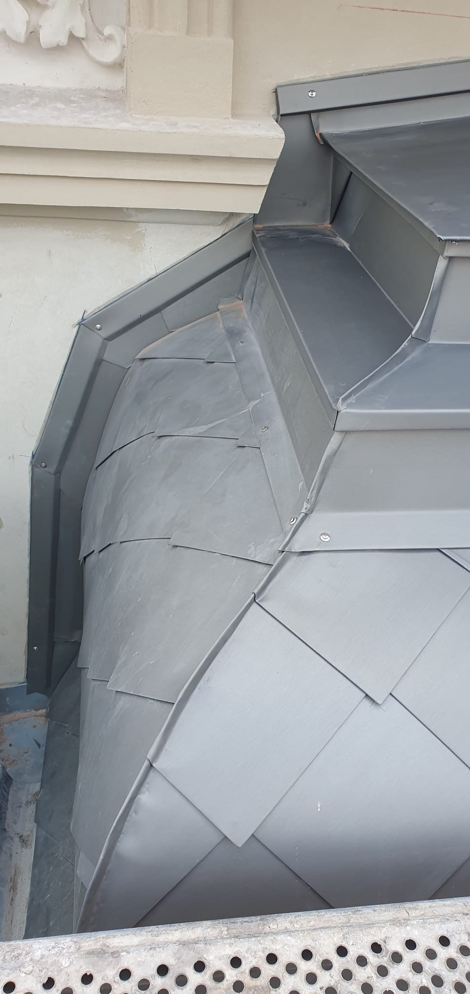 Szczegółowe ujęcie obróbki blacharskiej na dachu zabytkowej kamienicy, z widocznymi elementami dekoracyjnymi elewacji i metalowymi płytkami ułożonymi w unikalny wzór.