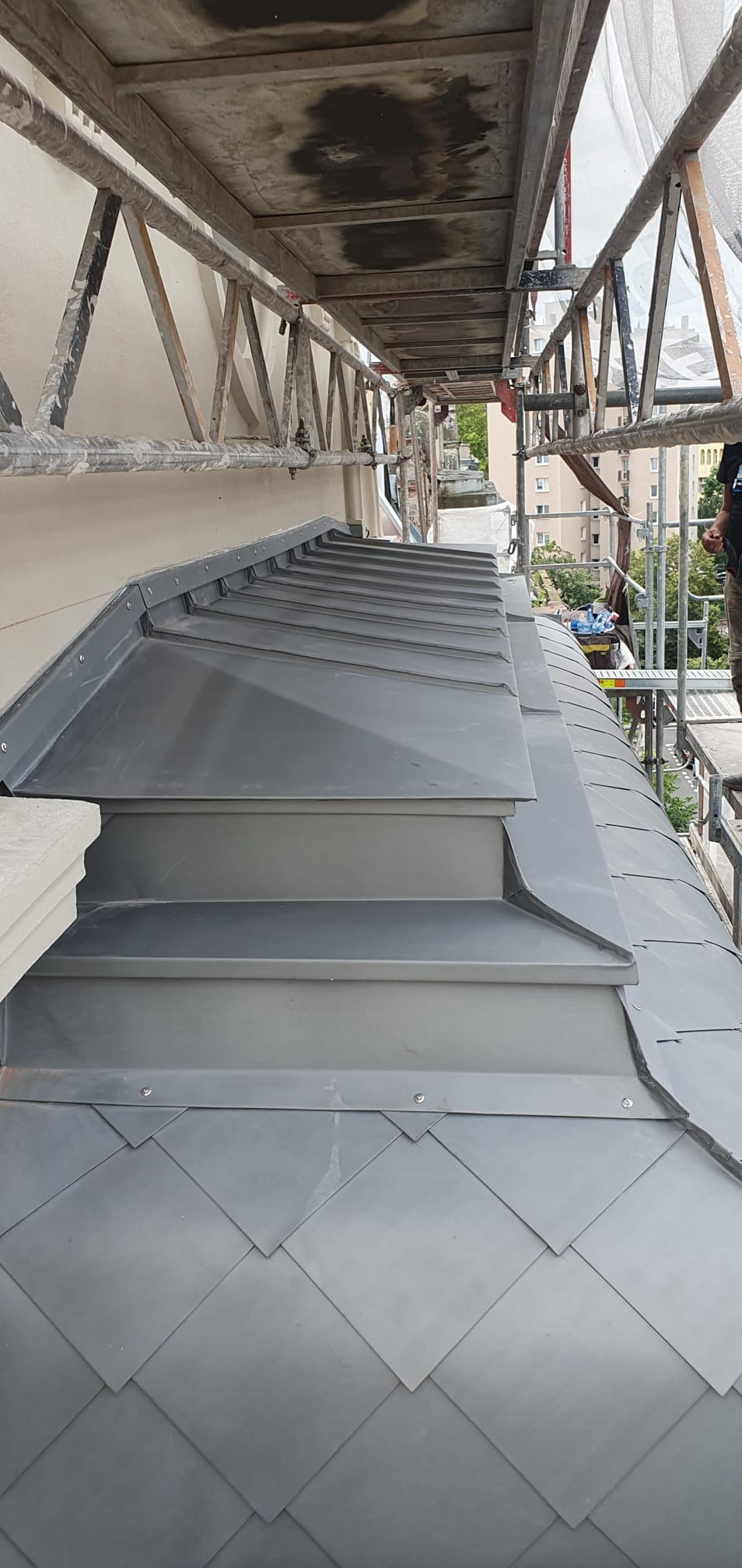 Szary dach z blachy łuskowej z lukarnami, widziany z rusztowania w Rogoźnie. Elementy mocowane na nity, widoczne detale obróbek.