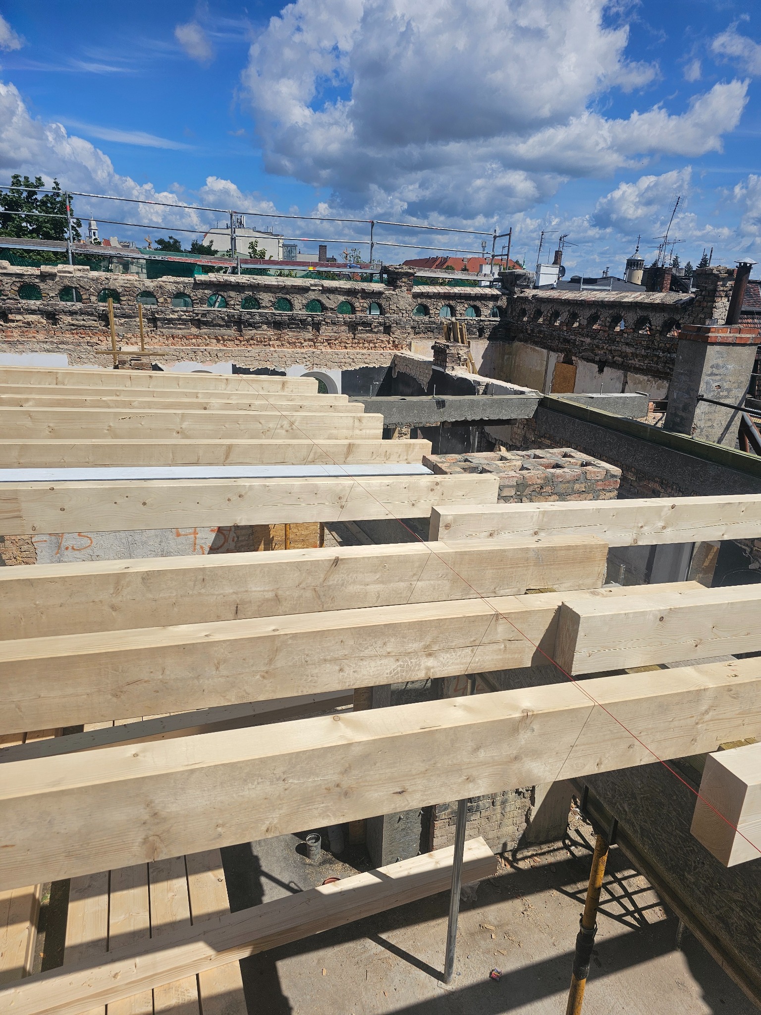 Wysoki kąt: Nowa więźba dachowa z jasnego drewna na tle starego, ceglanego budynku z widocznymi elementami zabezpieczeń i błękitnego nieba z chmurami.