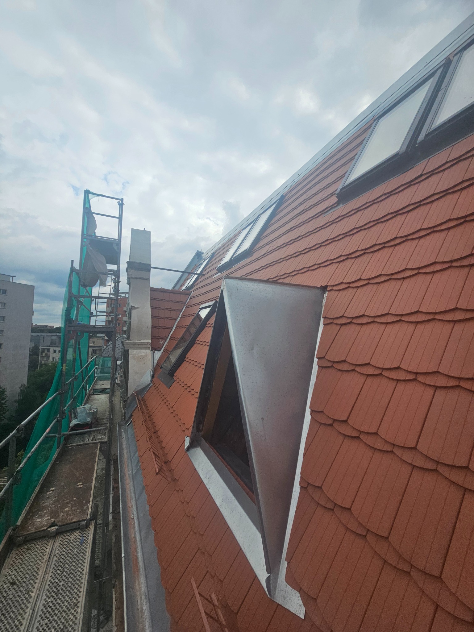 Czerwony dach z lukarnami i oknami dachowymi, widok z rusztowania. Elementy blacharskie i komin. Tło stanowi pochmurne niebo i budynki.