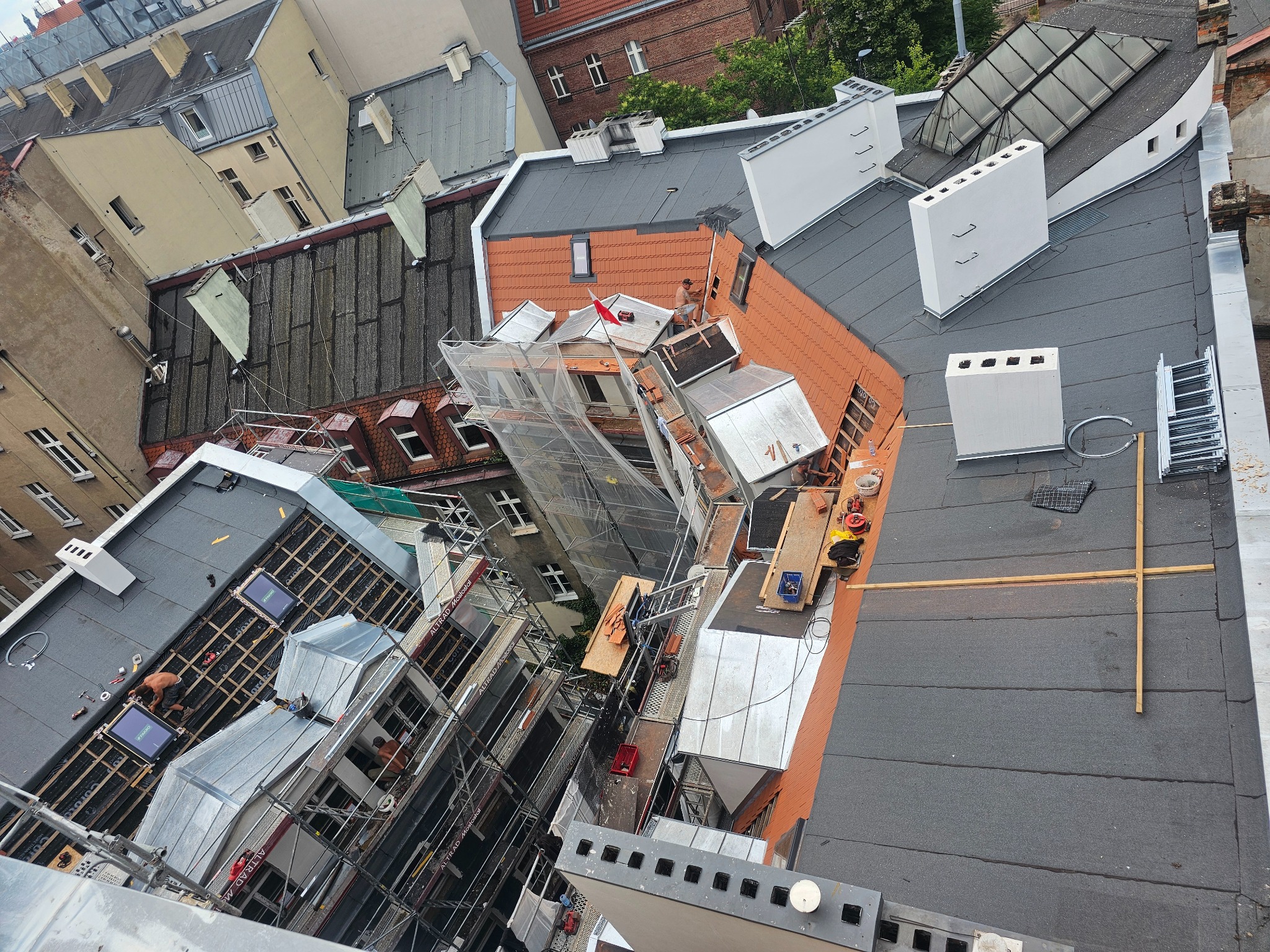 Prace dekarskie na dachu kamienicy z użyciem rusztowań. Widok z góry ukazuje fragmenty dachów w różnej fazie remontu i narzędzia pracy dekarzy.