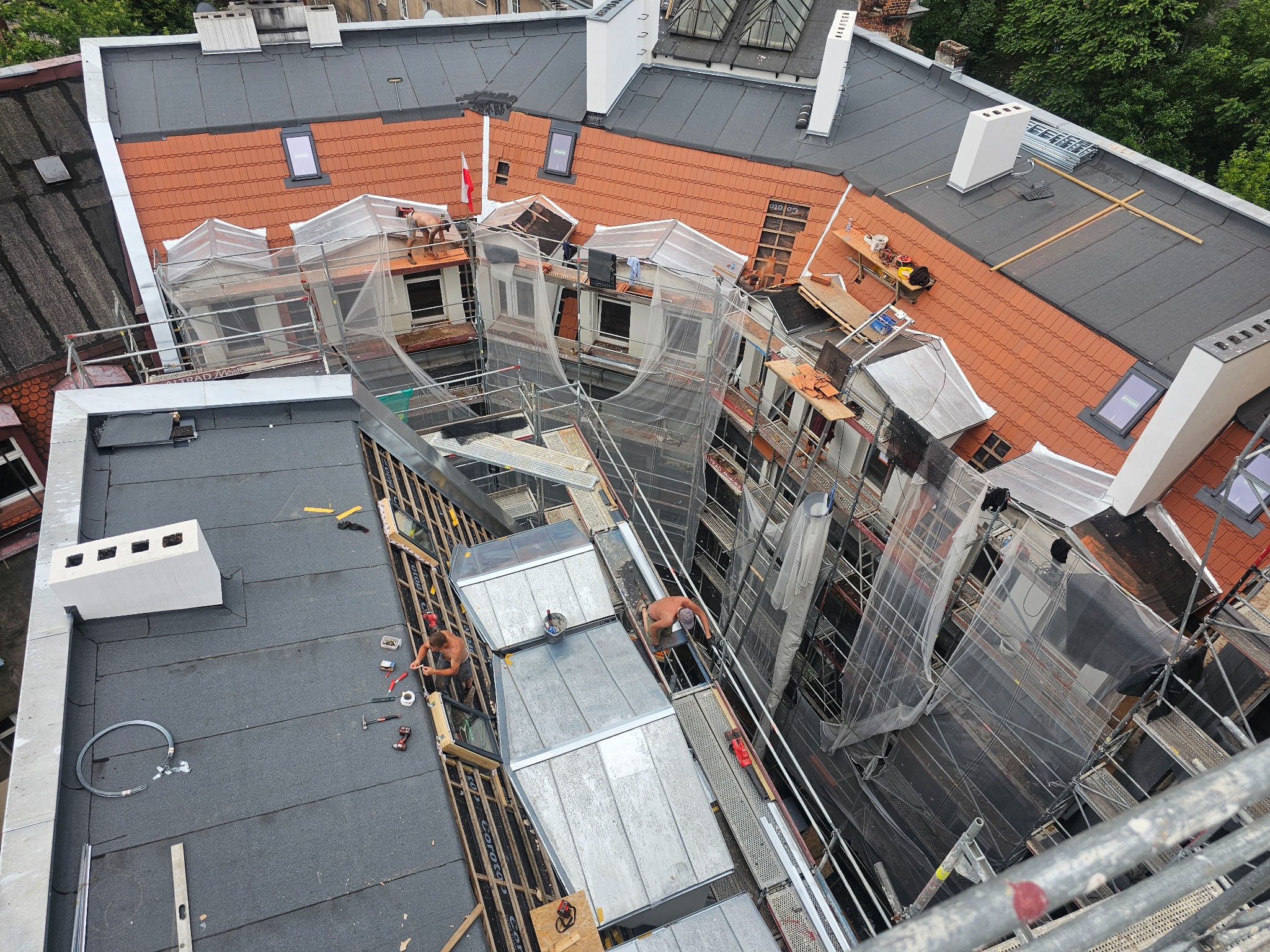 Widok z góry na budynek w trakcie prac dekarskich, z rusztowaniami i robotnikami na dachu. Różne rodzaje pokrycia dachowego i elementy konstrukcyjne w trakcie montażu.