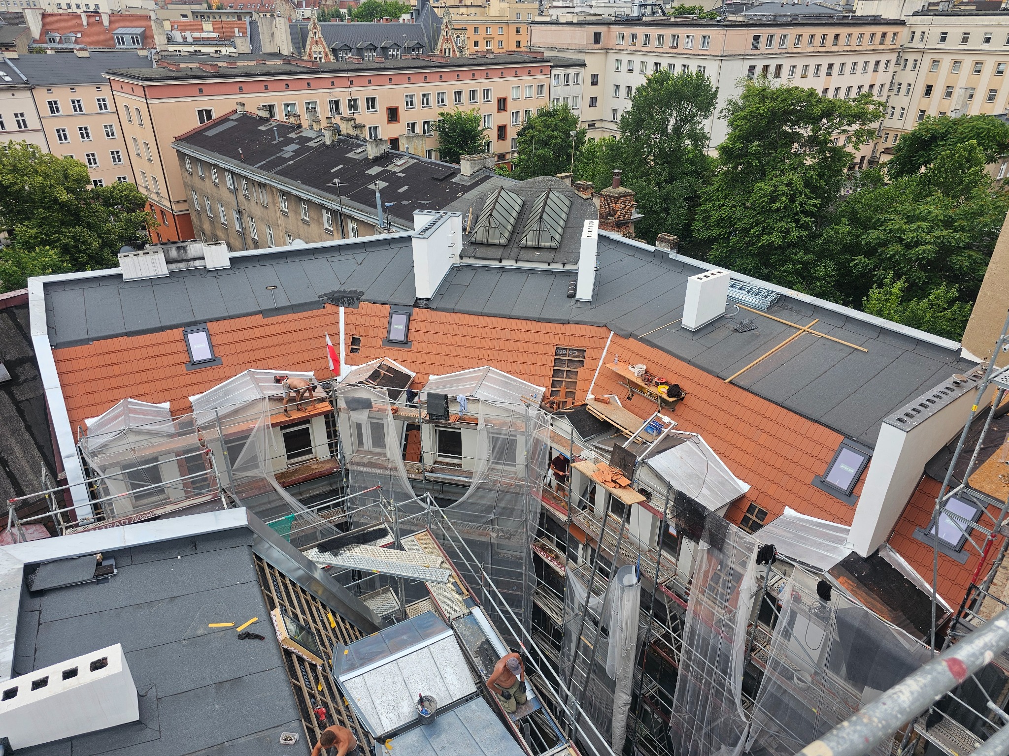 Widok z góry na budynek w trakcie renowacji. Część dachu pokryta czerwoną dachówką, część czarnym materiałem. Widoczne rusztowania i pracownicy. W tle zabudowa miejska.