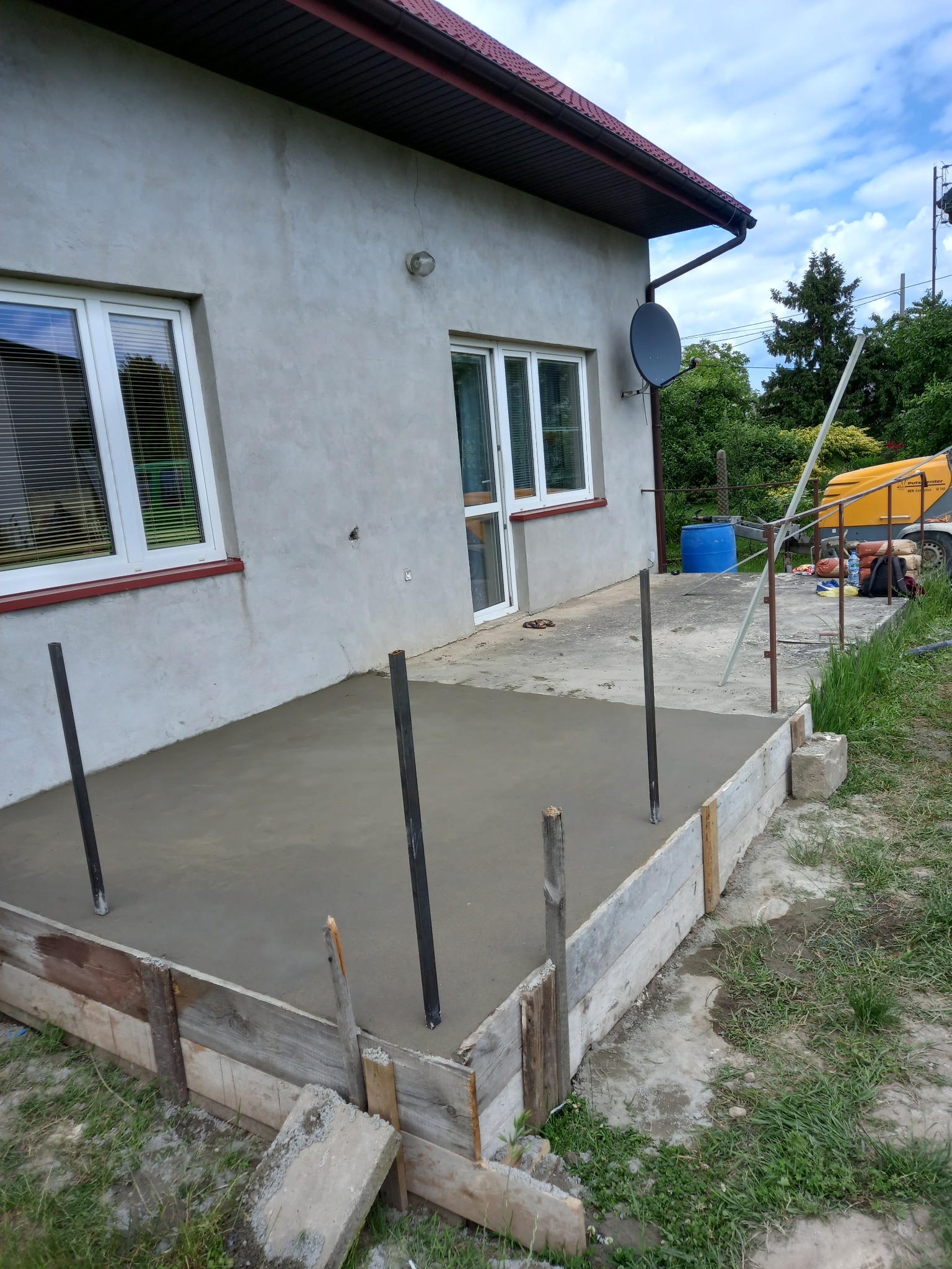 Świeżo wylana, gładka posadzka z jastrychu cementowego na zewnątrz budynku, z widocznym szalunkiem i wystającymi prętami zbrojeniowymi, w tle ściana domu i sprzęt budowlany.