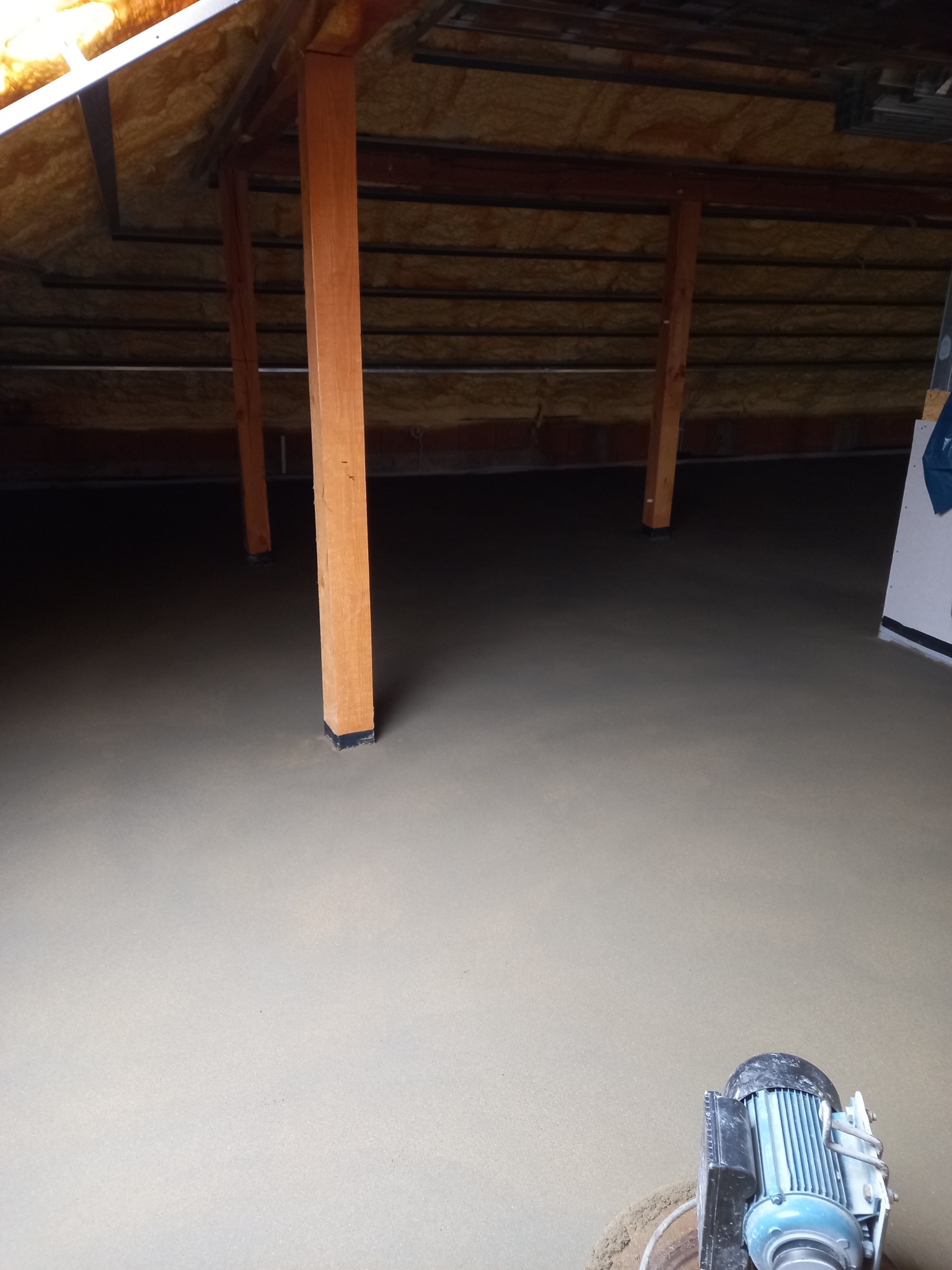 Świeżo wylana, jasnoszara posadzka w surowym wnętrzu poddasza, z widocznymi drewnianymi słupami podtrzymującymi konstrukcję dachu i maszyną do wygładzania posadzki w prawym dolnym rogu.