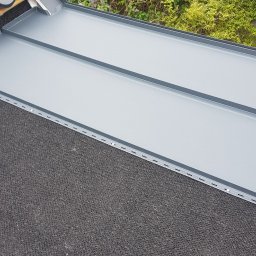 Dachnet - Szary, blaszany dach z widocznym fragmentem podbitki i bujną roślinnością w tle.