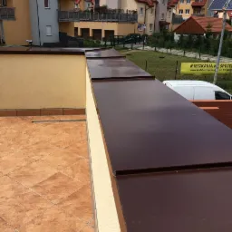 Nowoczesne brązowe obróbki blacharskie na dachu płaskim z terakotowymi płytkami w tle zabudowa wielorodzinna.