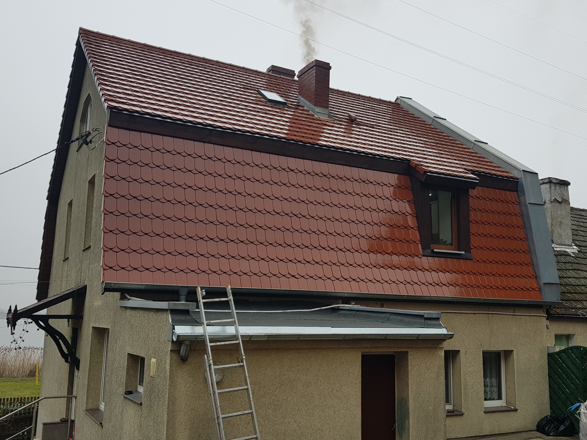 Dwupiętrowy dom z nowym, czerwonym dachem z dachówki ceramicznej, widoczna drabina oparta o rynnę, komin z dymem.