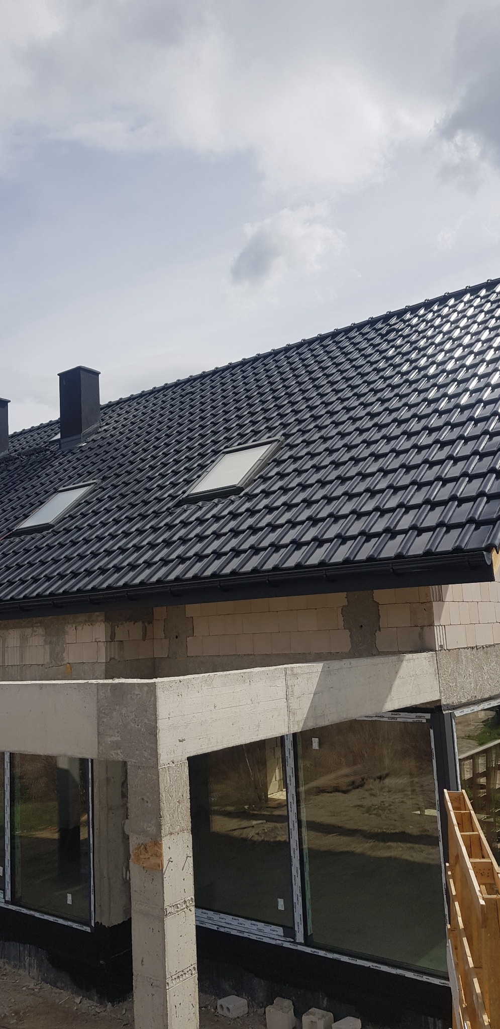 Nowy, ciemny dach z dwoma oknami dachowymi i czarnymi rynnami na surowym budynku z betonu komórkowego i dużymi oknami.