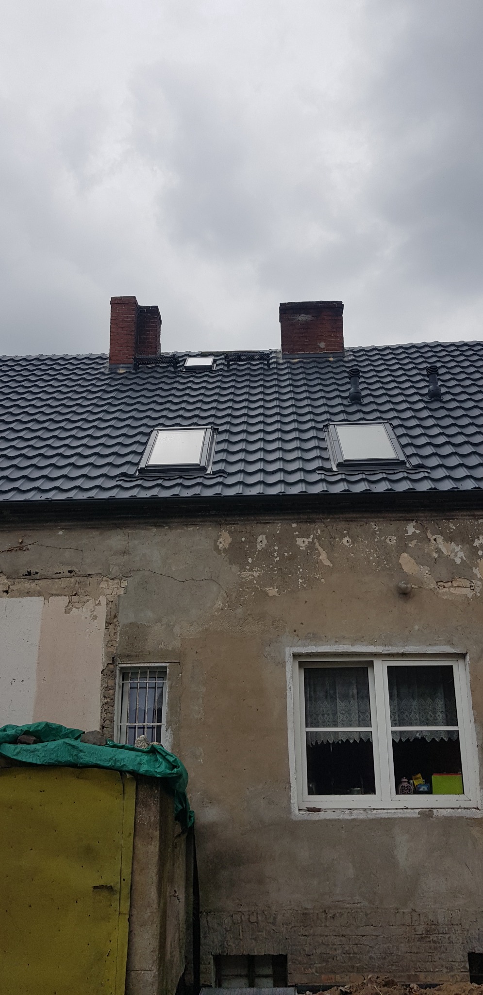 Ciemnoszary dach z dachówkami i oknami dachowymi na tle pochmurnego nieba, widoczny komin i fragment starego budynku z oknami.