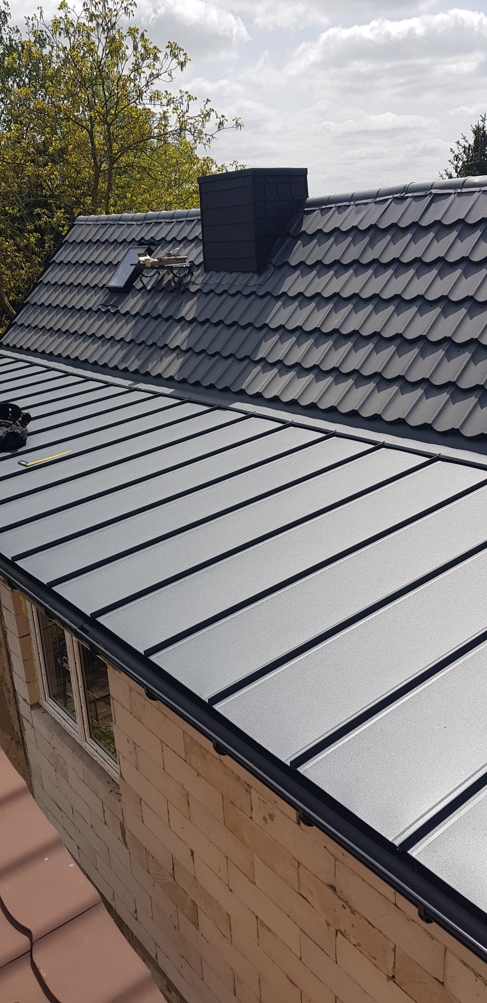 Szary dach z blachy trapezowej z kominem i oknem dachowym, widok z góry na budynek w trakcie budowy.