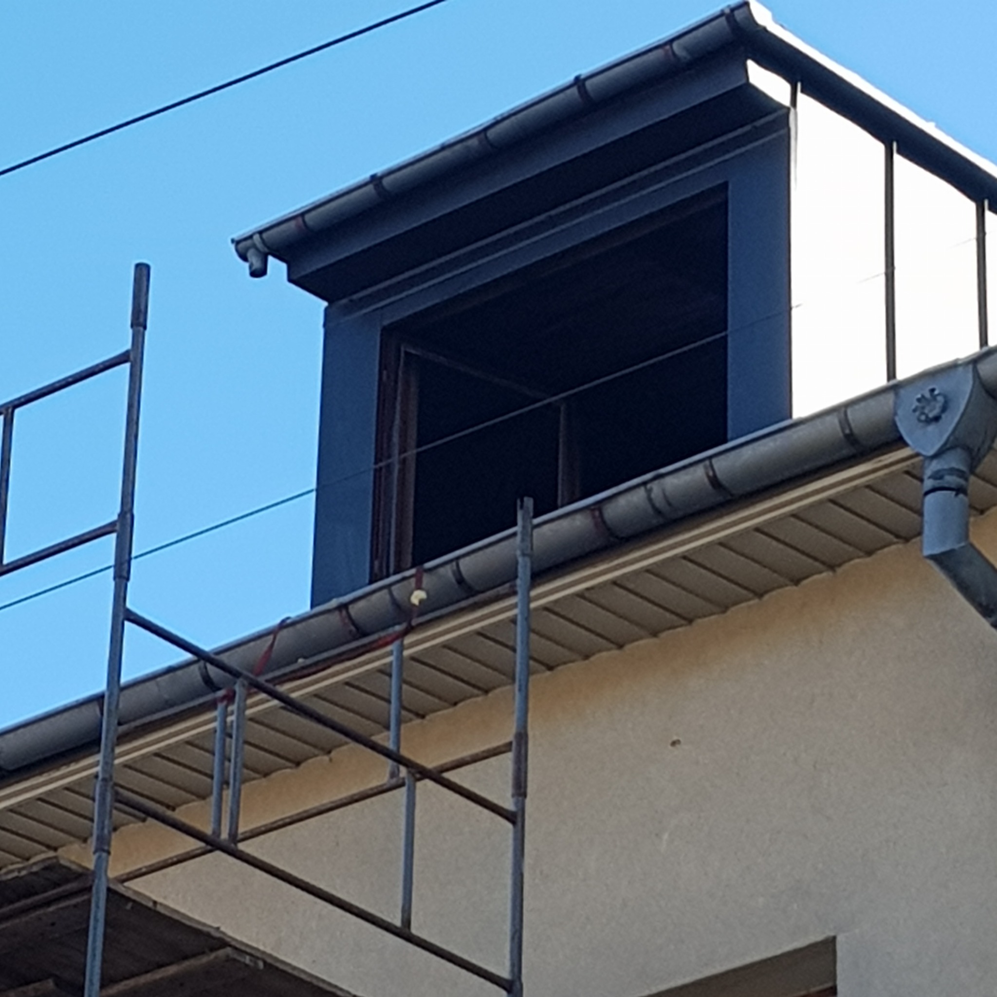 Fragment dachu z lukarną w trakcie budowy, widoczne rusztowanie i rynna z dekoracyjnym elementem.