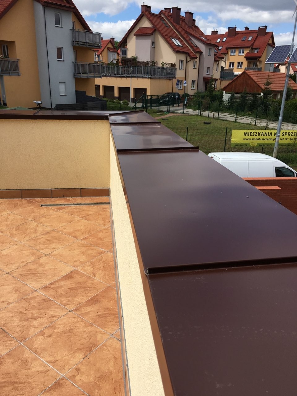 Nowoczesne brązowe obróbki blacharskie na dachu płaskim z terakotowymi płytkami w tle zabudowa wielorodzinna.