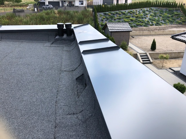 Widok z góry na płaski dach pokryty papą z dwoma kominkami wentylacyjnymi i obróbką blacharską w kolorze grafitowym, na tle zieleni ogrodowej i zabudowy jednorodzinnej.