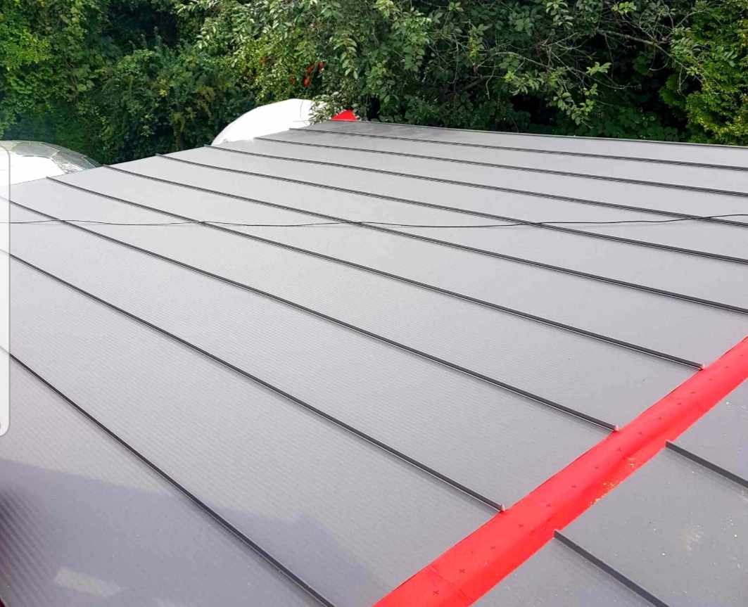 Szary dach z blachy na rąbek stojący, z widocznym czerwonym elementem uszczelniającym przy krawędzi, na tle zieleni drzew.