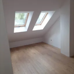 Poddasze po remoncie z dwoma oknami dachowymi i jasną podłogą z paneli, widoczny fragment ściany z niezakrytą puszką elektryczną.