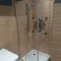 Narożna kabina prysznicowa z chromowanymi elementami i deszczownicą, w łazience wykończonej płytkami w odcieniach beżu z dekoracyjnym motywem kwiatowym, obok pralka.