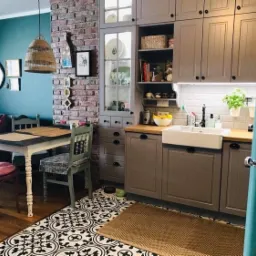 Kuchnia w stylu rustykalnym z ceglaną ścianą, szarymi szafkami, białą umywalką farmerską i turkusową lodówką, uzupełniona czarno-białą podłogą z wzorami i drewnianym stołem z krzesłami.