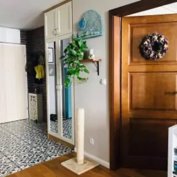 Przedpokój w stylu eklektycznym: białe drzwi wejściowe, wzorzysta podłoga, ceglana ściana, szafa z lustrem, drewniane drzwi wewnętrzne z wieńcem, roślina pnąca w szklanej kuli.