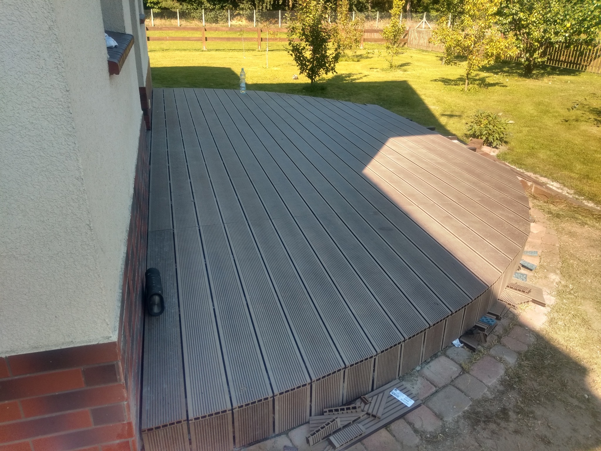 Kompozytowy taras przydomowy z zaokrąglonym wykończeniem, widok z góry w słoneczny dzień, z pozostawionymi fragmentami paneli na ziemi.