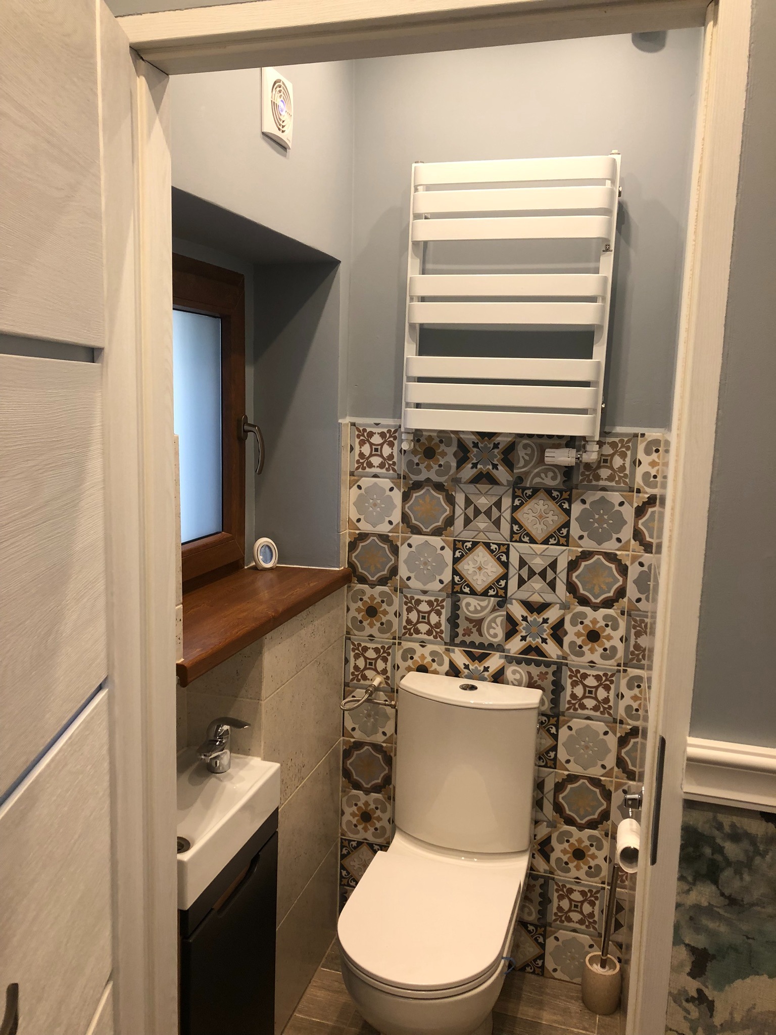 Niewielka łazienka po remoncie, z toaletą, umywalką w szafce, grzejnikiem drabinkowym, oknem z drewnianym parapetem i dekoracyjnymi płytkami na ścianie.
