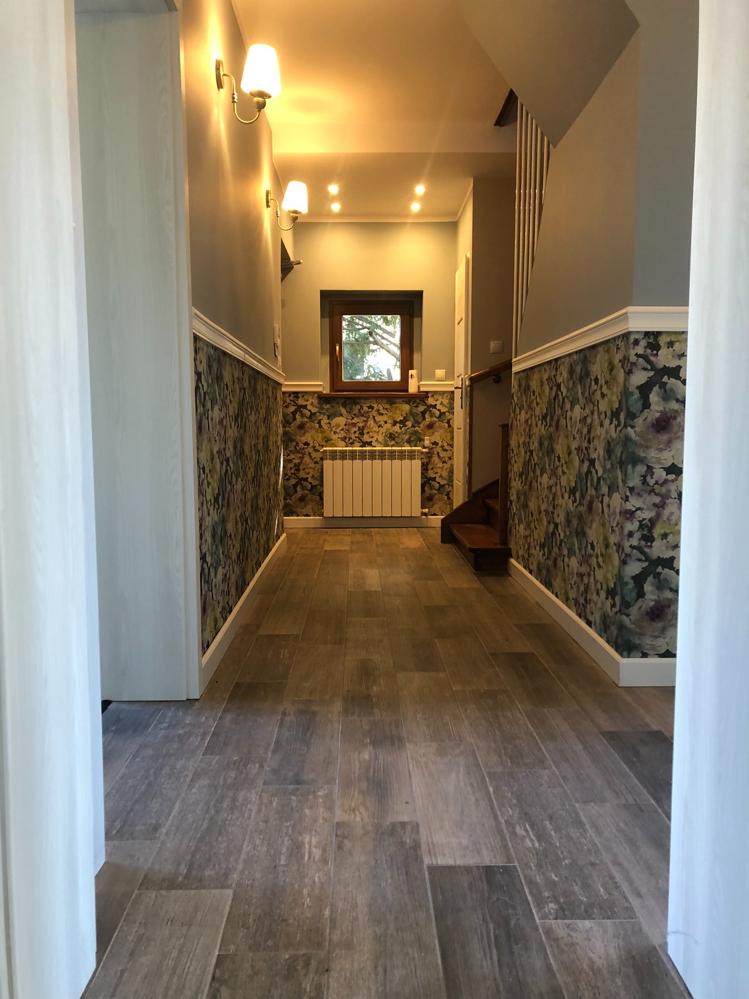 Korytarz po remoncie z panelami podłogowymi imitującymi drewno, ścianami w dolnej części wykończonymi tapetą w kwiaty i białymi listwami, oraz białymi, drewnianymi futrynami drzwi.