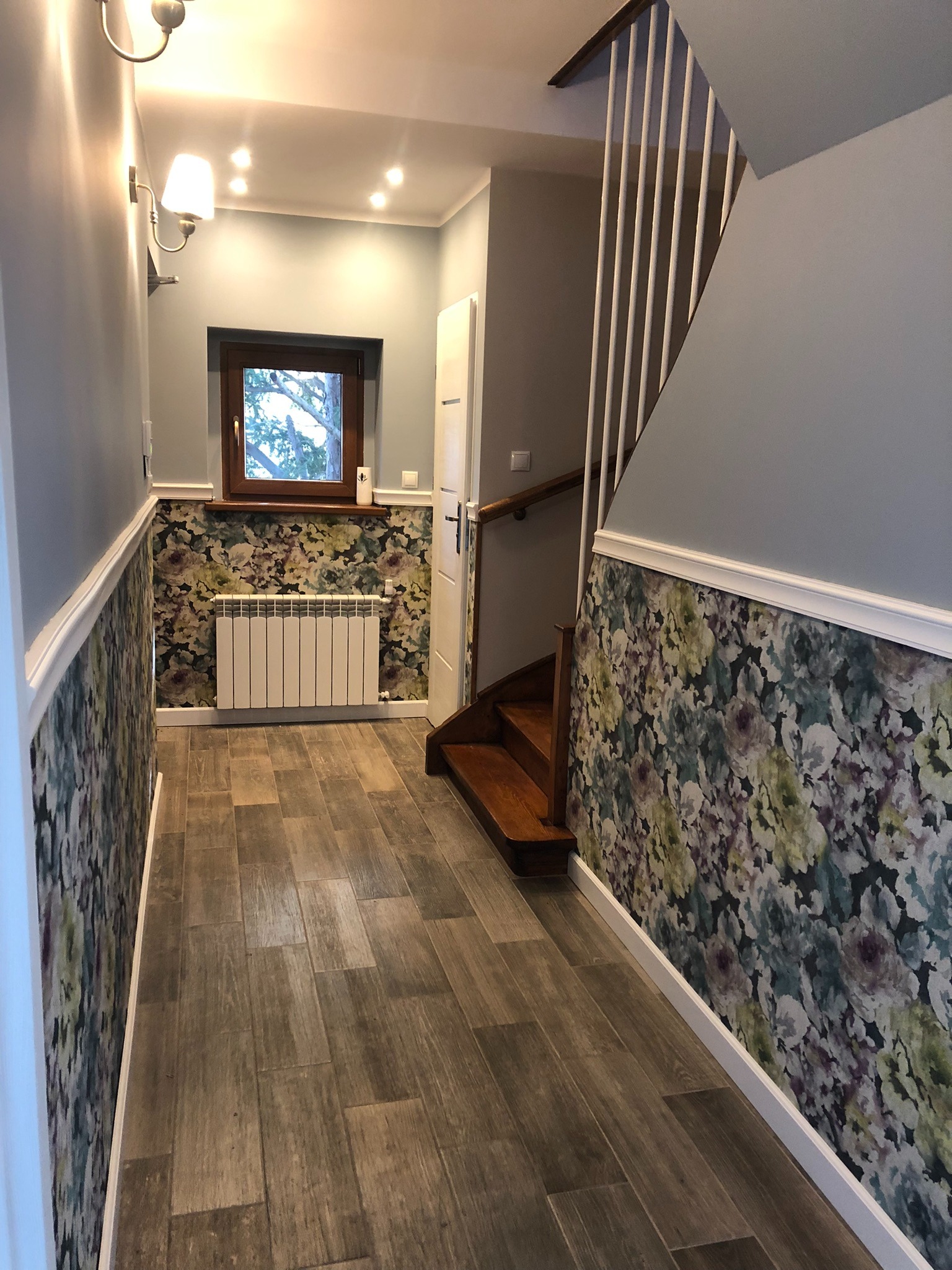 Korytarz po remoncie: drewnopodobna podłoga z paneli, ściany w dolnej części pokryte tapetą w kwiaty, w górnej części pomalowane na szaro, widoczne schody drewniane i biały grzejnik pod oknem.