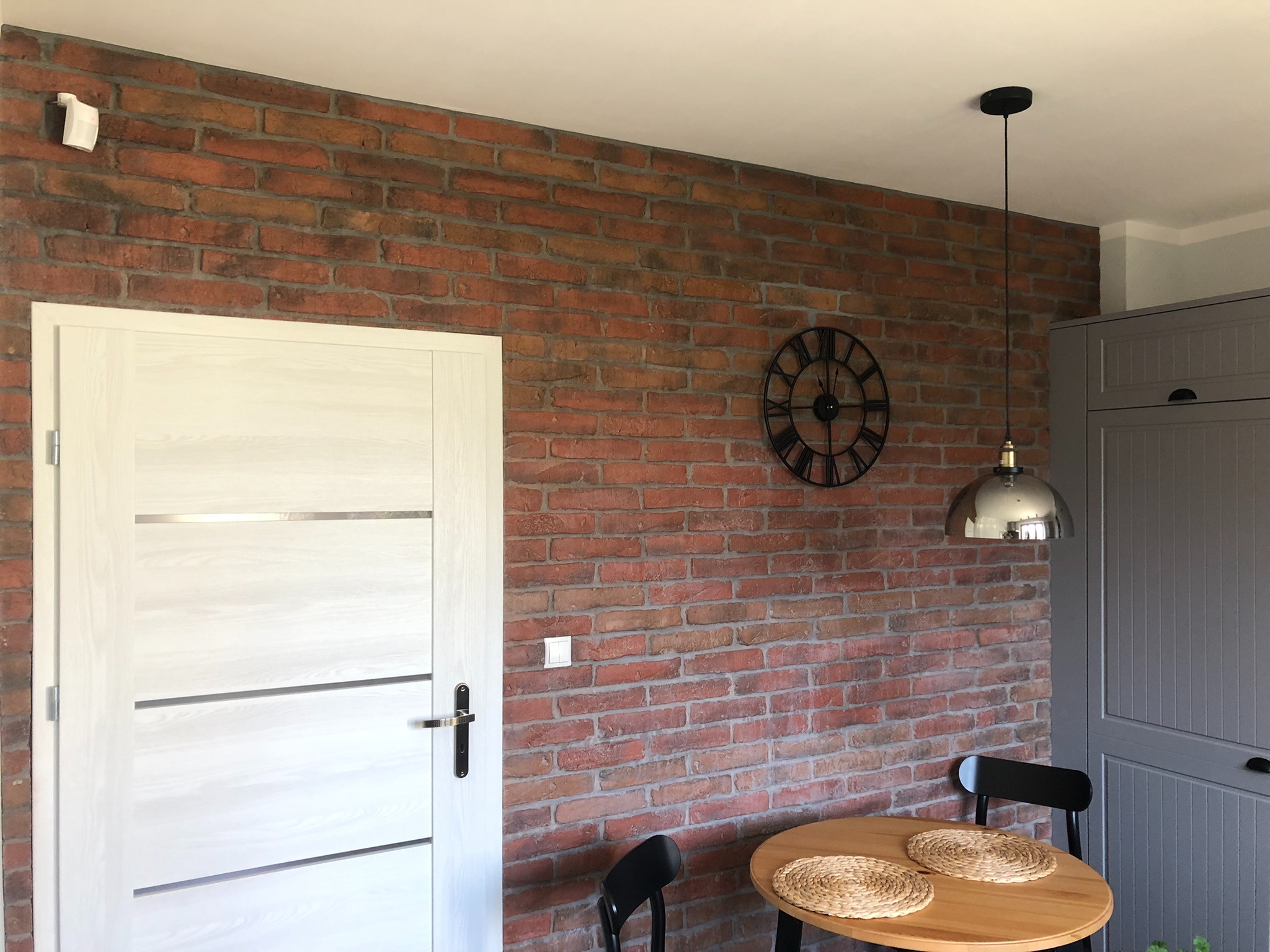 Wnętrze z dekoracyjną ścianą z czerwonej cegły, białe drzwi z poziomymi wstawkami, okrągły stół z plecionymi podkładkami, metalowa lampa wisząca oraz czarny zegar na ścianie.
