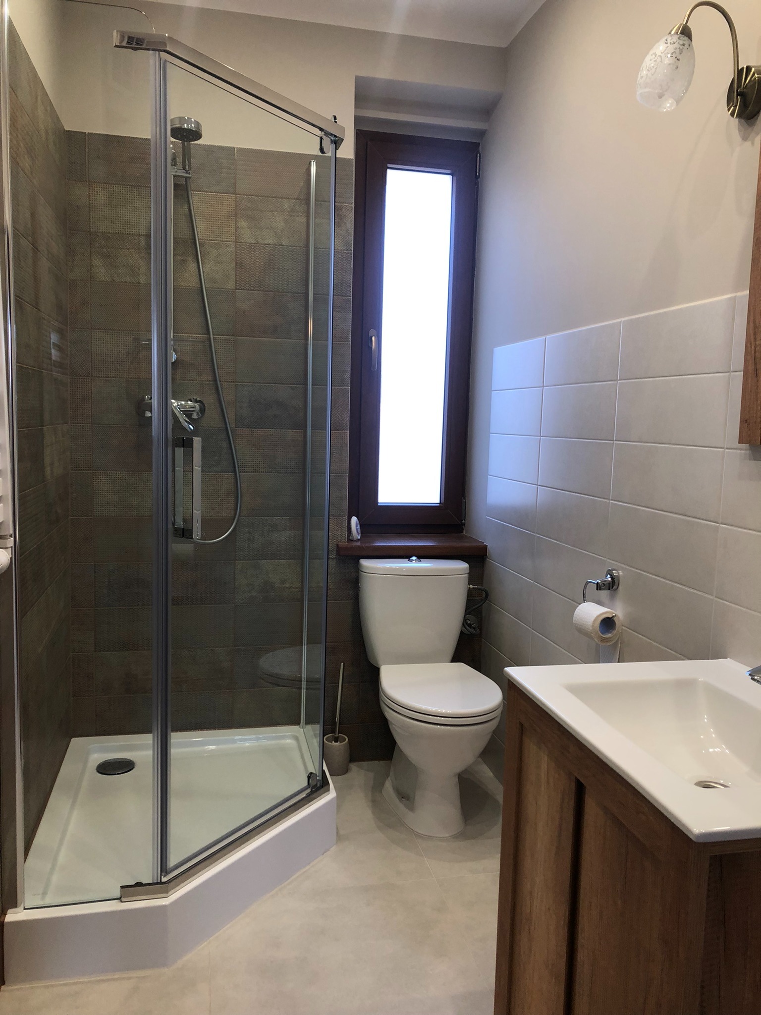 Nowoczesna łazienka z kabiną prysznicową narożną ze szklanymi drzwiami, toaletą pod oknem i umywalką z drewnianą szafką, wykończona płytkami w odcieniach brązu i beżu.