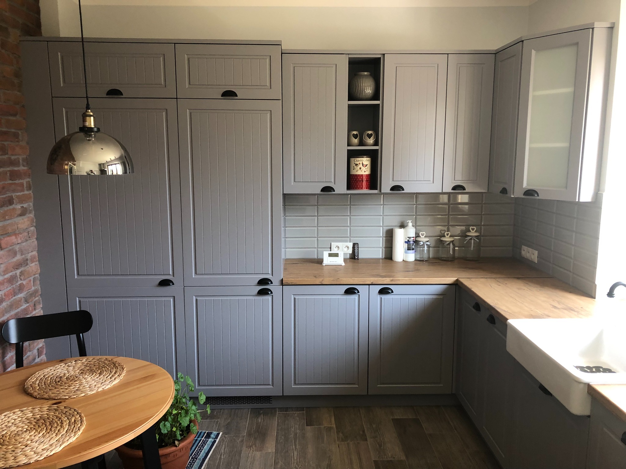 Szare meble kuchenne z drewnianym blatem i okładziną ścienną z szarych płytek, widoczny stół z plecionymi podkładkami i lampa w stylu retro.