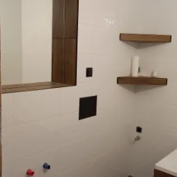 Nowoczesna łazienka z białymi płytkami, drewnianymi akcentami w postaci ramy lustra i półek, oraz podłogą imitującą drewno. Widoczne przygotowane przyłącza wodne.