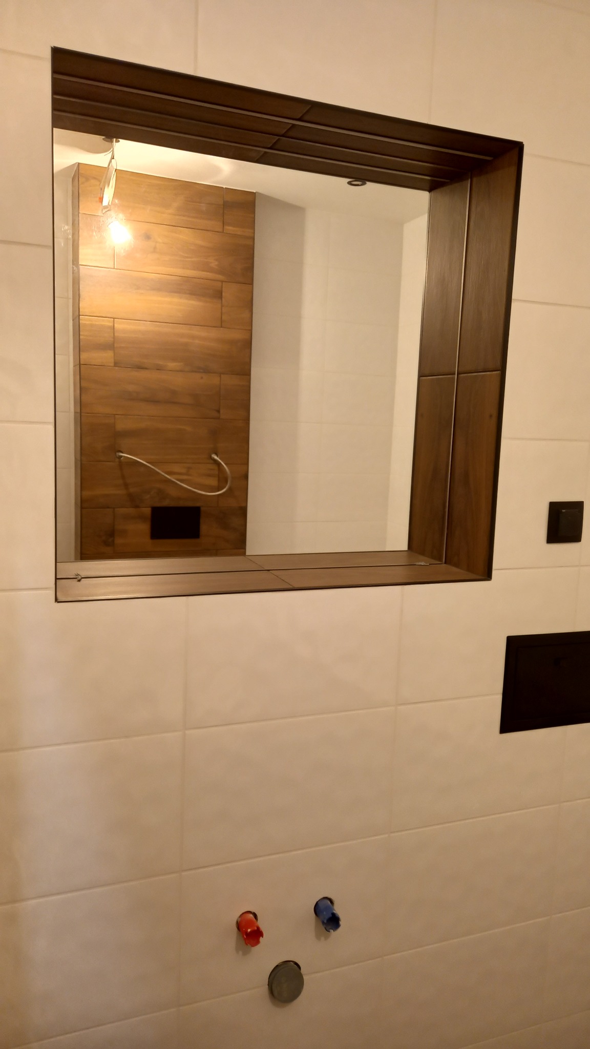 Wnęka z lustrem w ramie z ciemnego drewna na ścianie wyłożonej białymi płytkami. W lustrze odbija się ściana z drewnianymi panelami. Widoczne przyłącza wodne z czerwonymi i niebieskimi zaślepkami.