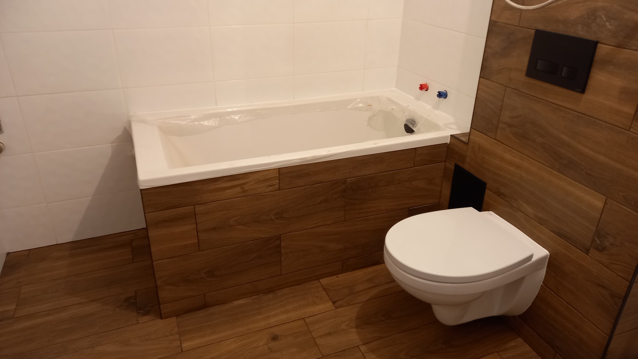 Nowoczesna łazienka w trakcie wykańczania: biała wanna zabezpieczona folią, podwieszana toaleta, brązowe płytki imitujące drewno na ścianach i podłodze, białe płytki na ścianie nad wanną, widoczne...