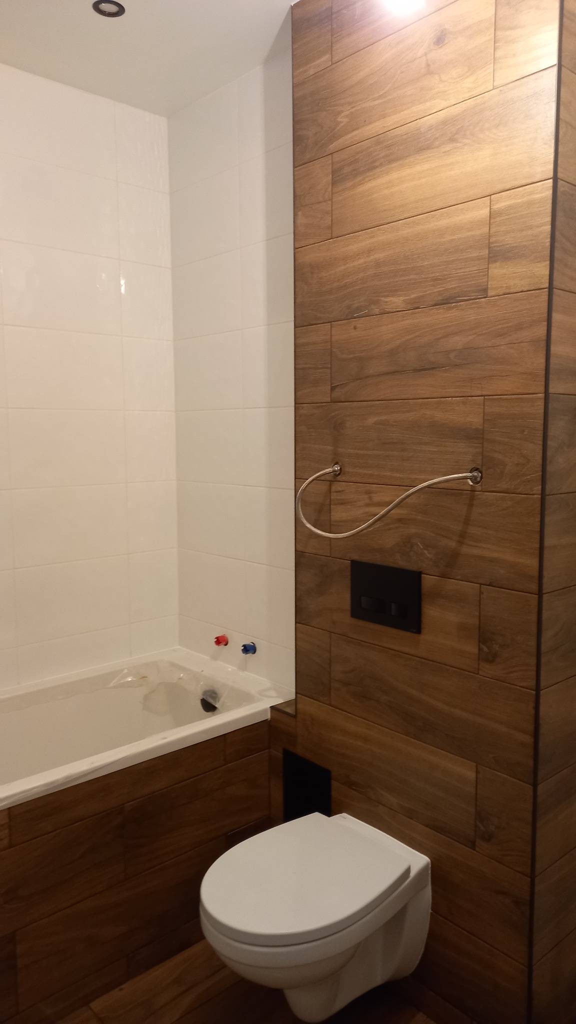 Nowoczesna łazienka z białymi płytkami i drewnopodobną okładziną, wisząca toaleta, bateria umywalkowa z czerwonym i niebieskim oznaczeniem, oraz metalowy wieszak na ręczniki.