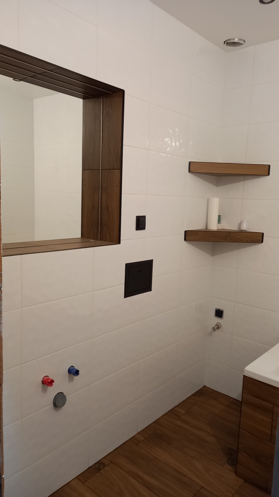 Nowoczesna łazienka z białymi płytkami, drewnianymi akcentami w postaci ramy lustra i półek, oraz podłogą imitującą drewno. Widoczne przygotowane przyłącza wodne.