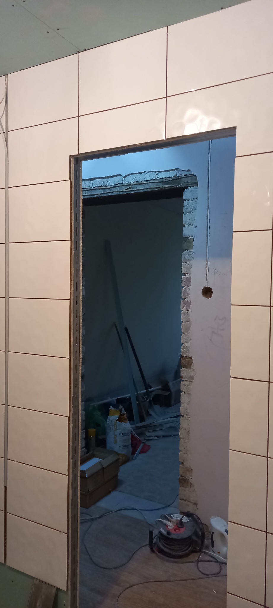 Białe płytki ułożone na ścianie w trakcie remontu łazienki, widoczny otwór drzwiowy z surową cegłą i wystającymi kablami elektrycznymi, w tle narzędzia i materiały budowlane.