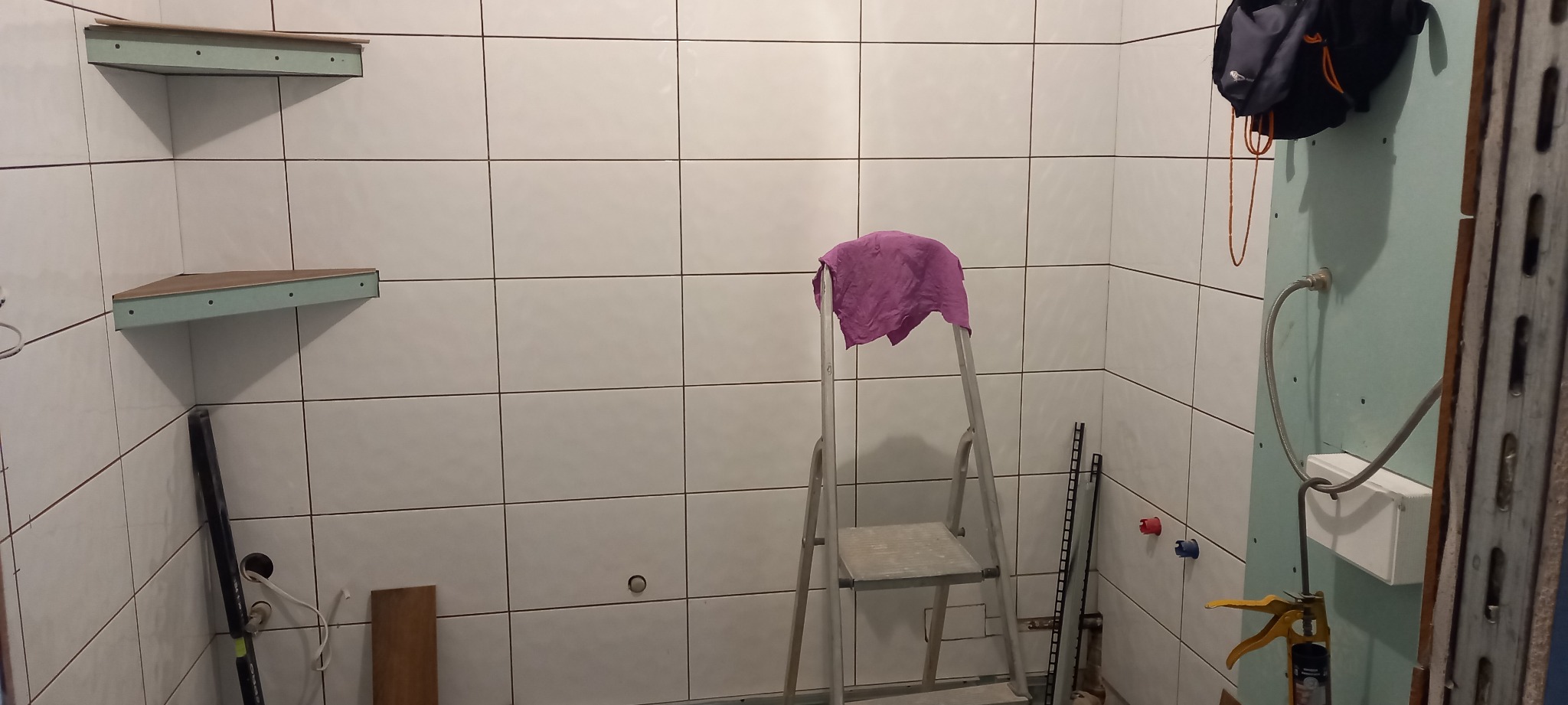 Wnętrze łazienki w trakcie prac: białe płytki, drabina z fioletowym ręcznikiem, zamontowane półki narożne z karton gipsu, fragment ściany z płyt karton gips z elementami instalacji.