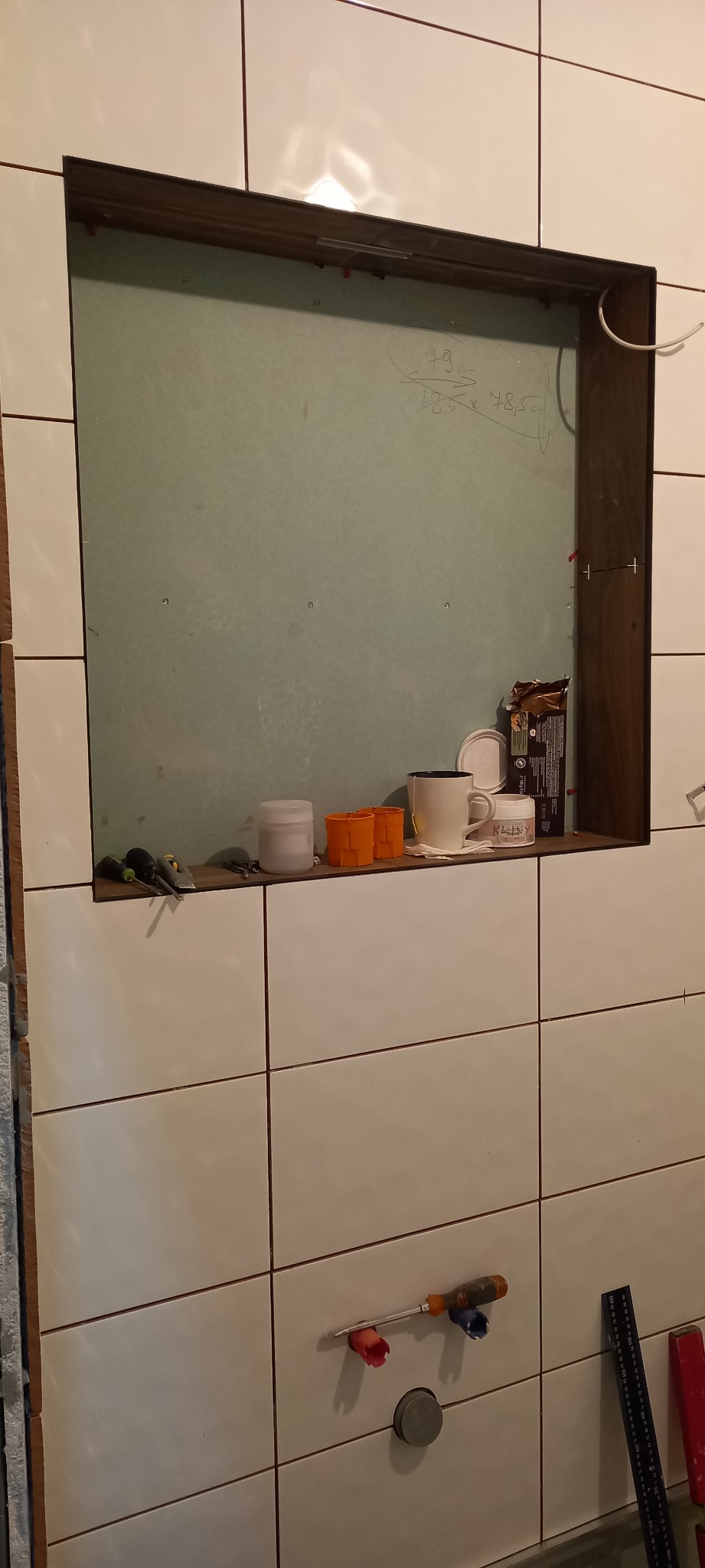 Niedokończona wnęka w łazience z płytą kartonowo-gipsową, na półce narzędzia, kubek i puszki, białe płytki na ścianie, widoczne ślady po wierceniu.