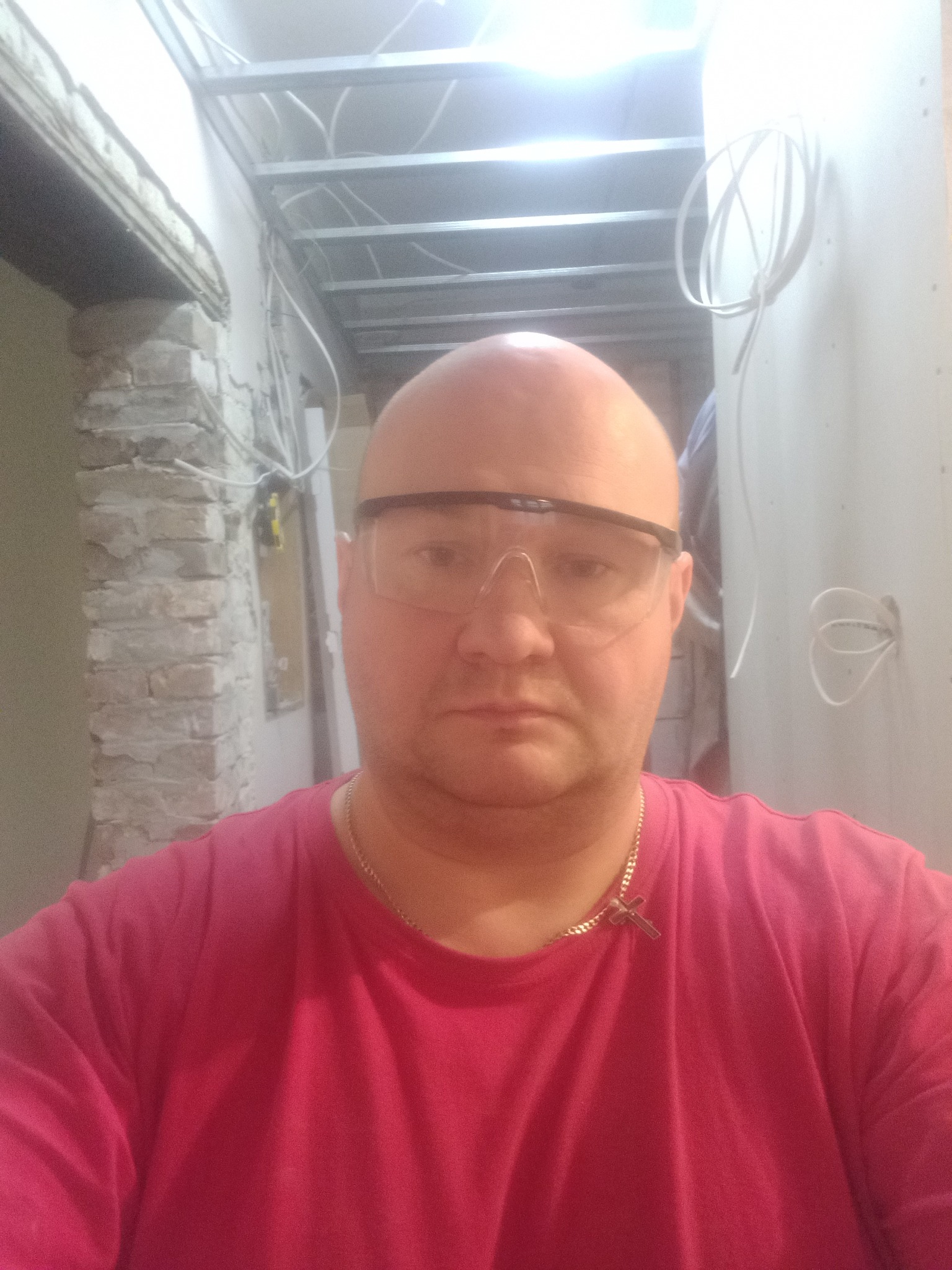 Mężczyzna w okularach ochronnych i różowej koszulce, w tle surowa ściana z instalacją elektryczną i metalową konstrukcją sufitu podwieszanego.