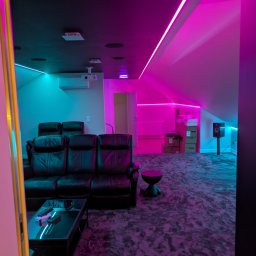 POWERDEK Klaudiusz Kulipczuk - Kino domowe w pokoju na poddaszu z czarną skórzaną sofą, stolikiem i oświetleniem LED w kolorach różu i turkusu. Widoczny projektor i głośnik.