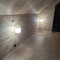 POWERDEK Klaudiusz Kulipczuk - Drewniane wnętrze poddasza z panelami ściennymi i podłogą w odcieniach beżu. Dwa kinkiety z wiklinowym wzorem oświetlają przestrzeń. Widoczne gniazdko elektryczne.