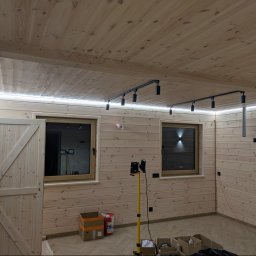 POWERDEK Klaudiusz Kulipczuk - Drewniane wnętrze z oświetleniem LED podsufitowym i reflektorami na szynoprzewodzie. Widoczne okna, drzwi stodołowe i narzędzia na podłodze.