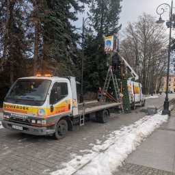 POWERDEK Klaudiusz Kulipczuk - Montaż dekoracji świątecznych na zewnątrz przez firmę POWERDEK. Pracownicy na podnośniku i drabinie dekorują choinkę. Na pierwszym planie samochód dostawczy firmy.