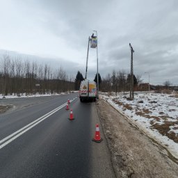 POWERDEK Klaudiusz Kulipczuk - Pracownik w podnośniku koszowym naprawia oświetlenie uliczne. Na drodze ustawione pachołki ostrzegawcze, w tle las i pochmurne niebo. Widoczny samochód serwisowy.