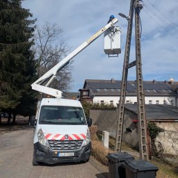 POWERDEK Klaudiusz Kulipczuk - Pracownik w podnośniku koszowym przy betonowym słupie energetycznym. Biały samochód z podnośnikiem na tle zabudowań i drzew. Dwie czarne beczki na śmieci na pierwszym planie.