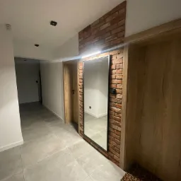 Nowoczesny korytarz z dużym lustrem w czarnej ramie, obudowanym cegłą, nad którym zamontowano podłużną lampę LED. Szare płytki podłogowe i drewniane drzwi w tle.