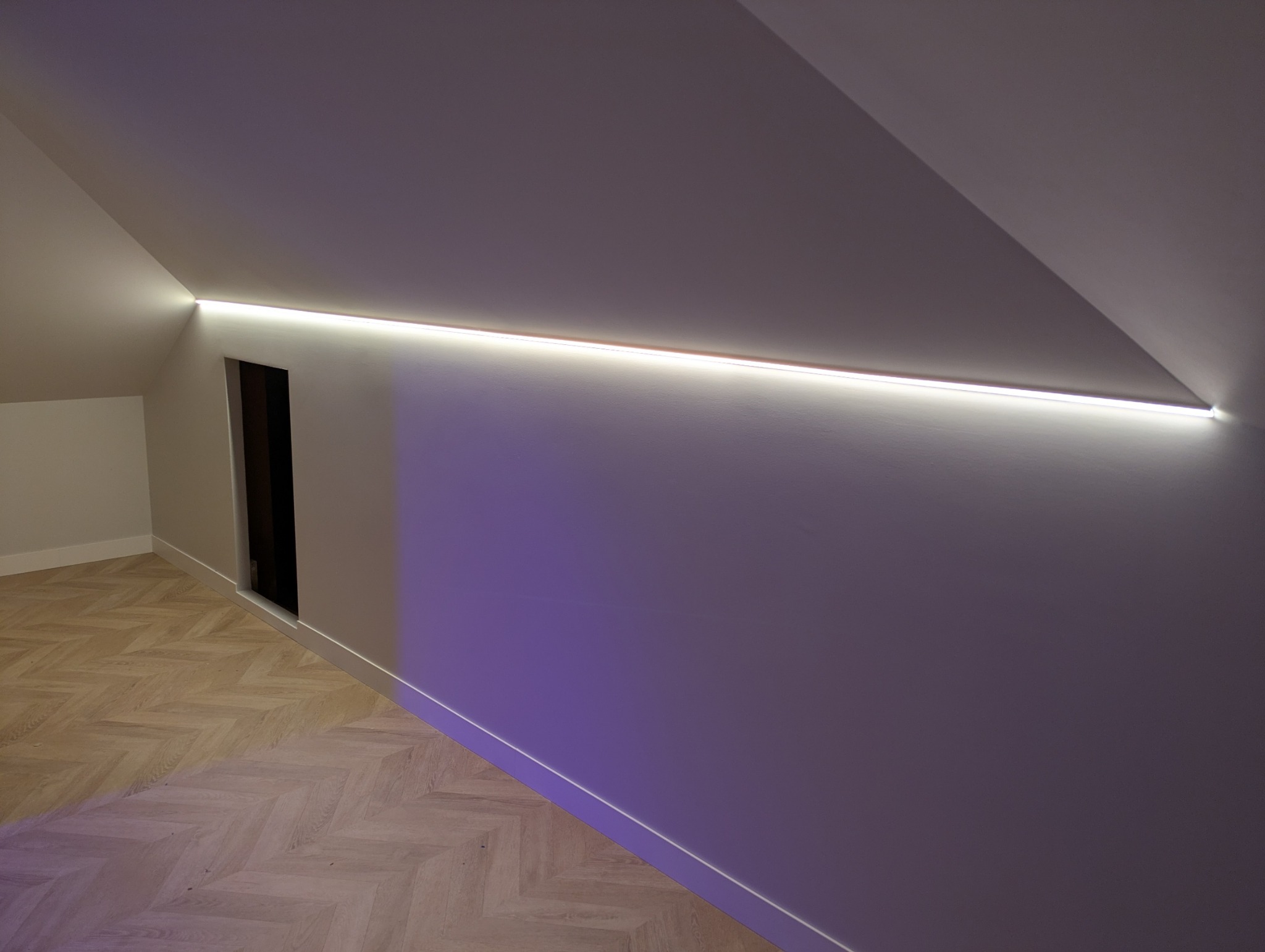 Oświetlenie LED wkomponowane w skos sufitu poddasza, z widocznym wejściem do garderoby i parkietem ułożonym w jodełkę. Nowoczesny design z subtelnym fioletowym podświetleniem ściany.